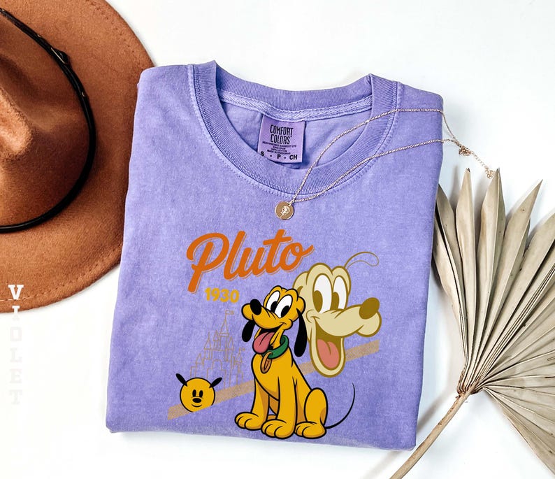 Może przedstawiać: Liliowa koszulka Comfort Colors z grafiką Plutona, psa z kresk&oacute;wek Disneya, oraz napisem "Pluto 1930". Projekt zawiera kresk&oacute;wkowy wizerunek Plutona i mały obraz twarzy Plutona. Złoty naszyjnik jest zarzucony na koszulkę.