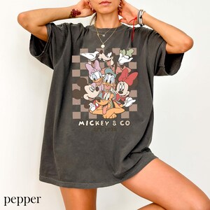 May include: Dark grey t-shirt with a graphic of Mickey Mouse and friends, including Minnie, Donald, Daisy, and Goofy, on a checkered background. The text "MICKEY & CO. EST. 1928" is printed below the characters. The shirt has short sleeves.