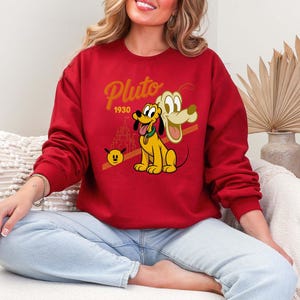 Może przedstawiać: Czerwona bluza z postacią z kresk&oacute;wki Pluto. Projekt zawiera napis "Pluto" złotym pismem, rok "1930" oraz wizerunek Plutona z sylwetką zamku. Bluza jest noszona przez osobę.