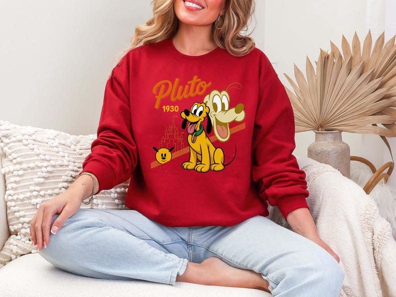 Może przedstawiać: Czerwona bluza z postacią z kresk&oacute;wki Pluto. Projekt zawiera napis "Pluto" złotym pismem, rok "1930" oraz wizerunek Plutona z sylwetką zamku. Bluza jest noszona przez osobę.
