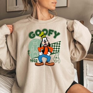 Może przedstawiać: Beżowa bluza z nadrukiem Goofy'ego. Grafika przedstawia Goofy'ego w czapce kucharskiej, pomarańczowej koszuli, niebieskich spodniach i brązowych butach. Słowo "GOOFY" jest zielone, a pod nim "1932". Po prawej stronie zielono-biała szachownica.