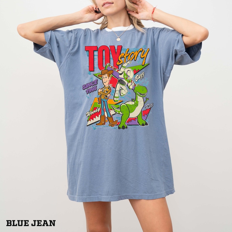 May include: Blue jean colored oversized t-shirt featuring a Toy Story graphic. The graphic includes Woody, Buzz Lightyear, and Rex, with the text "Toy Story Since 1995". The shirt has a white collar.