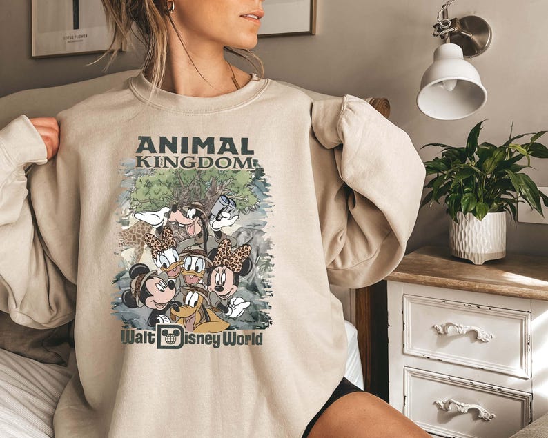 May include: Beige sweatshirt featuring a graphic of Disney characters, including Mickey and Minnie Mouse, Donald and Daisy Duck, and Goofy. The text "ANIMAL KINGDOM" and "Walt Disney World" are printed on the front. The sweatshirt has long sleeves.