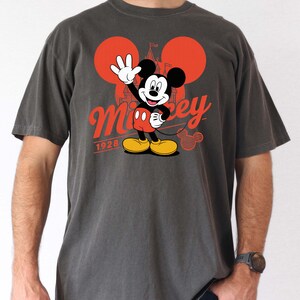 Może przedstawiać: Ciemnoszara koszulka z grafiką Myszki Miki. Projekt przedstawia Myszka Miki machającego, w czerwonych spodenkach i ż&oacute;łtych butach. Słowo "Mickey" jest w czerwonym skrypcie, z napisem "1928" poniżej. Dwa czerwone koła i sylwetka zamku dopełniają projekt.