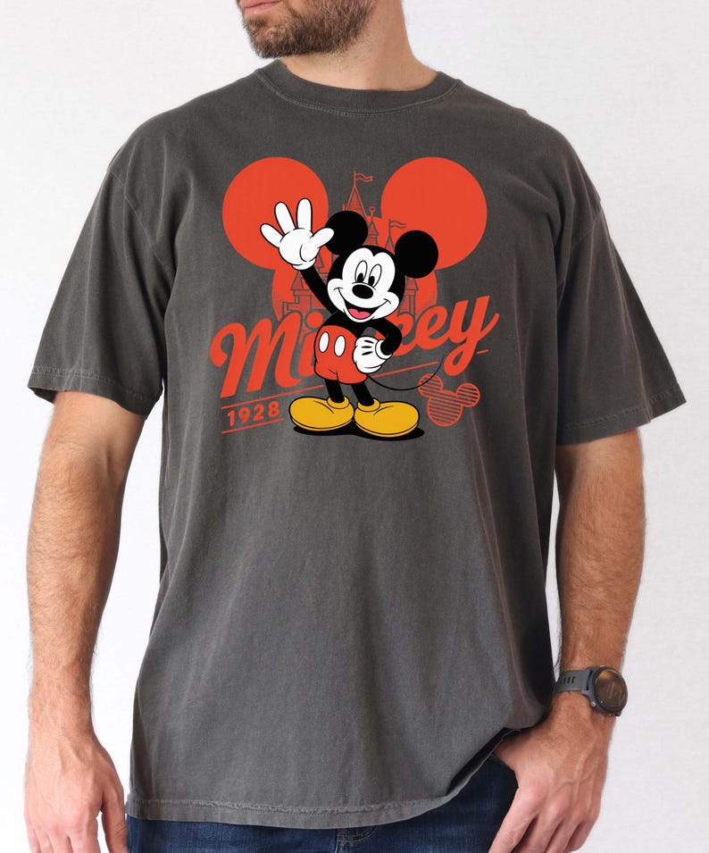 Może przedstawiać: Ciemnoszara koszulka z grafiką Myszki Miki. Projekt przedstawia Myszka Miki machającego, w czerwonych spodenkach i ż&oacute;łtych butach. Słowo "Mickey" jest w czerwonym skrypcie, z napisem "1928" poniżej. Dwa czerwone koła i sylwetka zamku dopełniają projekt.