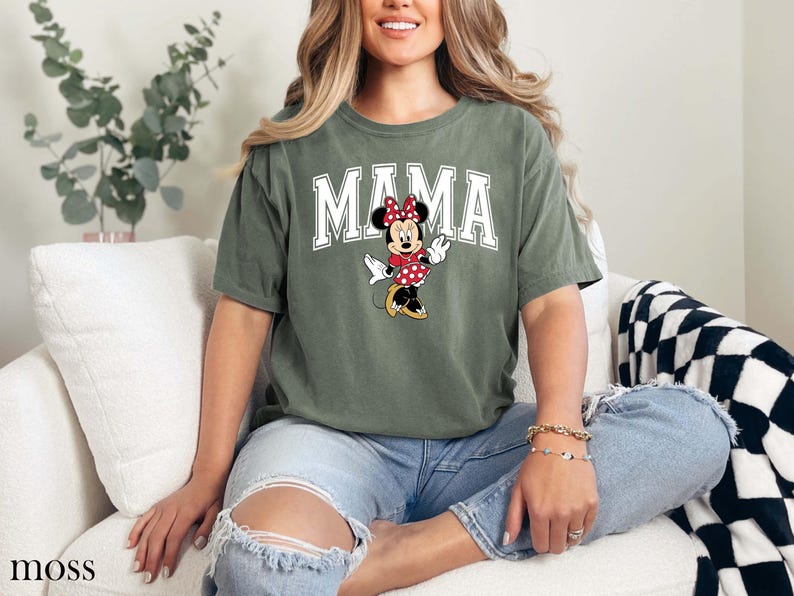 May include: Moss green t-shirt with the word "MAMA" in white block letters and a Minnie Mouse graphic. The graphic features Minnie Mouse wearing a red polka dot bow and dress. The shirt is a casual style.