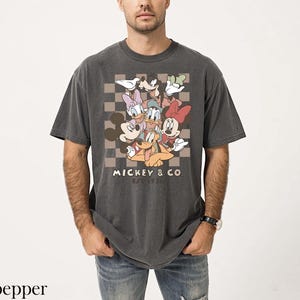 May include: Dark grey t-shirt with a graphic of Mickey Mouse and friends, including Minnie, Donald, and Daisy Duck, on a checkered background. The text "Mickey & Co. Est. 1928" is printed below the characters. The shirt is worn by a person.