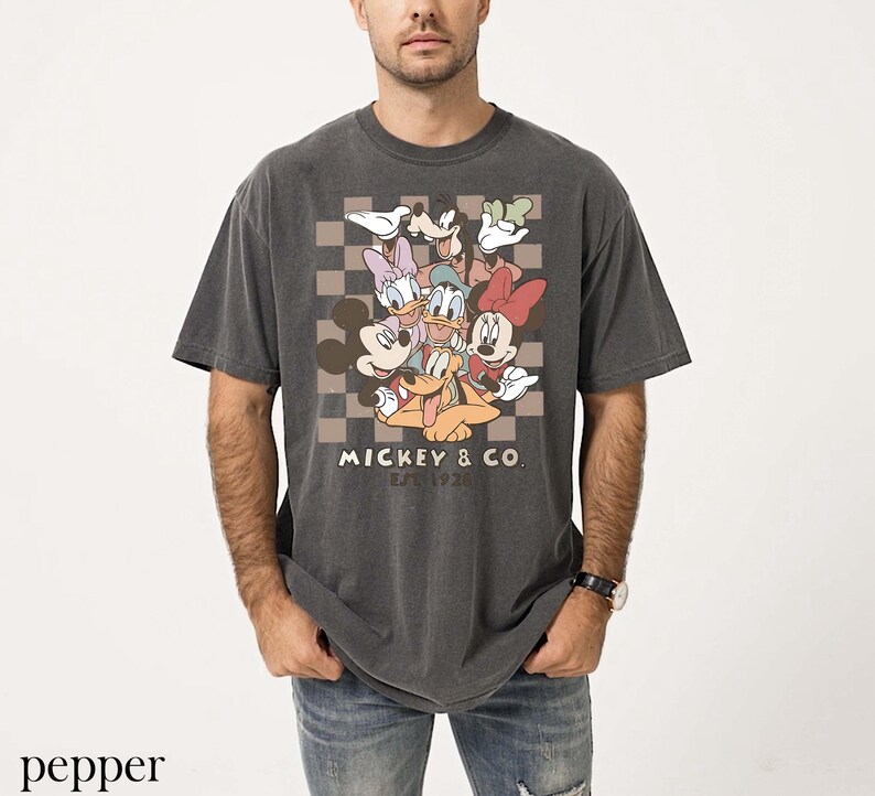 May include: Dark grey t-shirt with a graphic of Mickey Mouse and friends, including Minnie, Donald, and Daisy Duck, on a checkered background. The text "Mickey & Co. Est. 1928" is printed below the characters. The shirt is worn by a person.