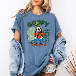 Może przedstawiać: Niebieska koszulka z Goofym. Projekt zawiera zielony napis "GOOFY", rok "1932" oraz wizerunek Goofy'ego w jego klasycznym stroju. Obecny jest r&oacute;wnież mały kontur Goofy'ego.