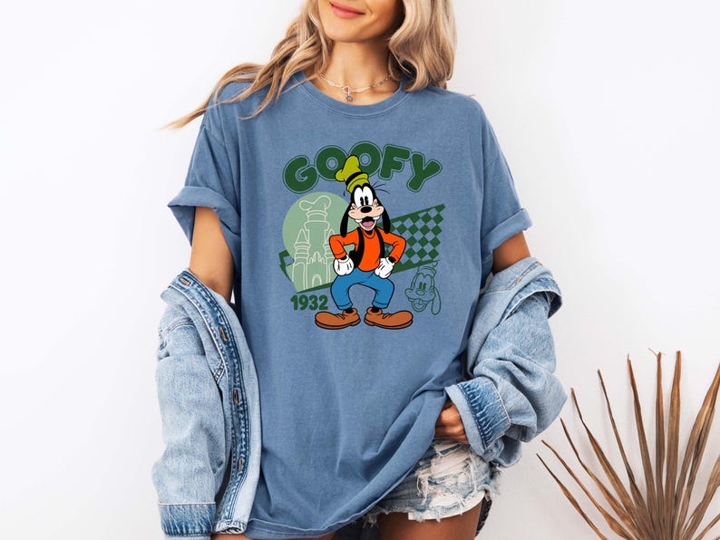 Może przedstawiać: Niebieska koszulka z Goofym. Projekt zawiera zielony napis "GOOFY", rok "1932" oraz wizerunek Goofy'ego w jego klasycznym stroju. Obecny jest r&oacute;wnież mały kontur Goofy'ego.