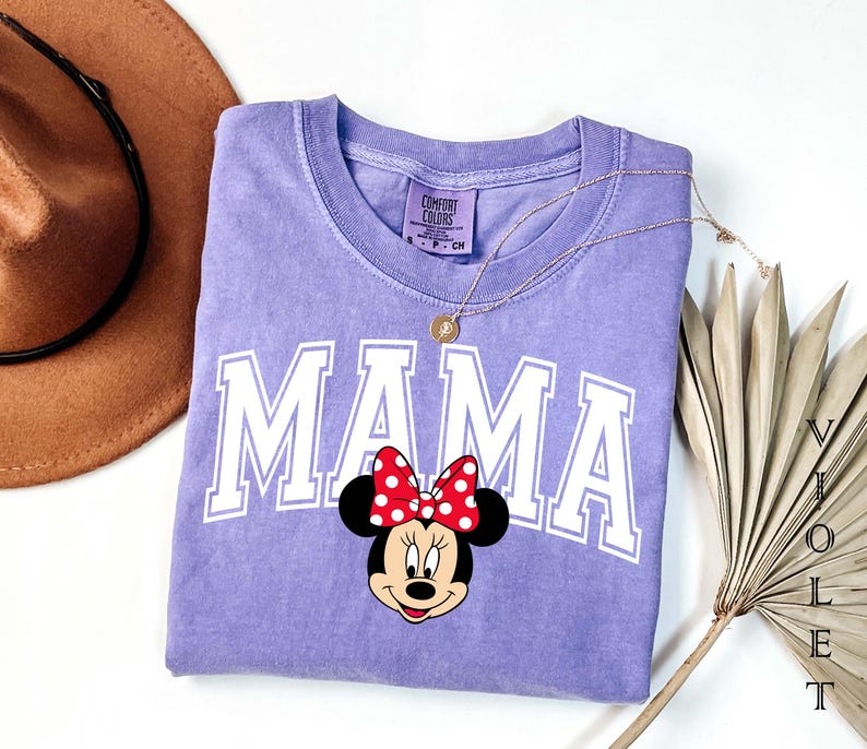 May include: A lilac Comfort Colors t-shirt with the word "MAMA" in white block letters and a Minnie Mouse graphic. The Minnie Mouse graphic has a red bow with white polka dots. A gold necklace and a brown hat are also in the image.