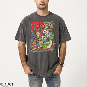 May include: A charcoal gray t-shirt featuring a graphic from the movie Toy Story. The design includes Woody, Buzz Lightyear, and Rex, with the text "TOY STORY" and "SINCE 1995". The shirt is a vintage style, with a slightly faded appearance.