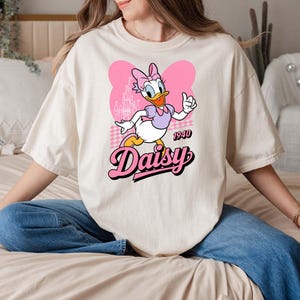 Może przedstawiać: Kremowa koszulka z kresk&oacute;wkowym wizerunkiem Daisy Duck. Daisy ma na sobie fioletowy top i r&oacute;żową kokardę, z napisem "Daisy" w r&oacute;żowej czcionce retro. Koszulka zawiera r&oacute;wnież rok "1940".