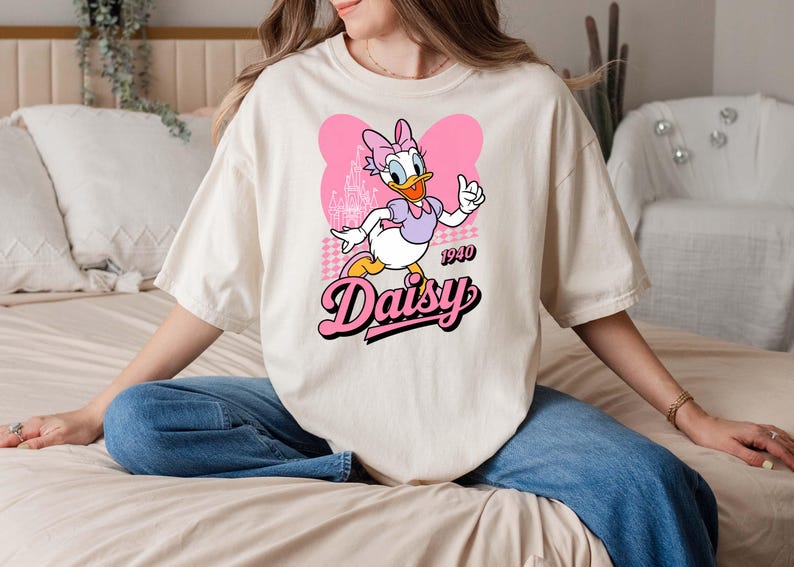 Może przedstawiać: Kremowa koszulka z kresk&oacute;wkowym wizerunkiem Daisy Duck. Daisy ma na sobie fioletowy top i r&oacute;żową kokardę, z napisem "Daisy" w r&oacute;żowej czcionce retro. Koszulka zawiera r&oacute;wnież rok "1940".