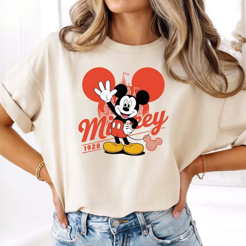 Może przedstawiać: Kremowa koszulka z grafiką Myszki Miki machającej, z czerwonymi akcentami i napisem "Mickey" w stylu vintage. Rok "1928" jest wydrukowany poniżej. Projekt zawiera kultowe uszy Myszki Miki i sylwetkę zamku.