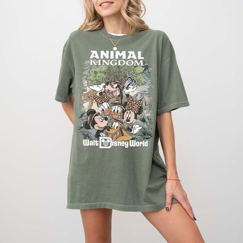 May include: Olive green t-shirt featuring a graphic of Disney characters, including Mickey and Minnie Mouse, Donald Duck, and Goofy. The shirt has the text "ANIMAL KINGDOM" and "Walt Disney World" in white.