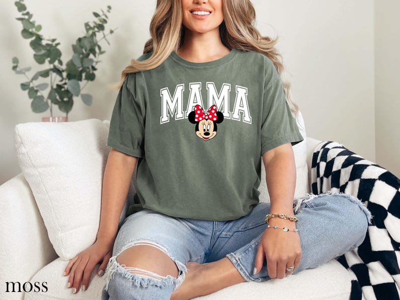 May include: A moss green t-shirt with the word "MAMA" in white block letters and a Minnie Mouse graphic. The shirt is a relaxed fit and has short sleeves. The shirt is made of a soft material.