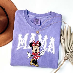 May include: A light purple t-shirt with the word "MAMA" in white block letters and a Minnie Mouse graphic. Minnie is wearing a red polka dot dress and bow. A gold necklace and a brown hat are also in the image.