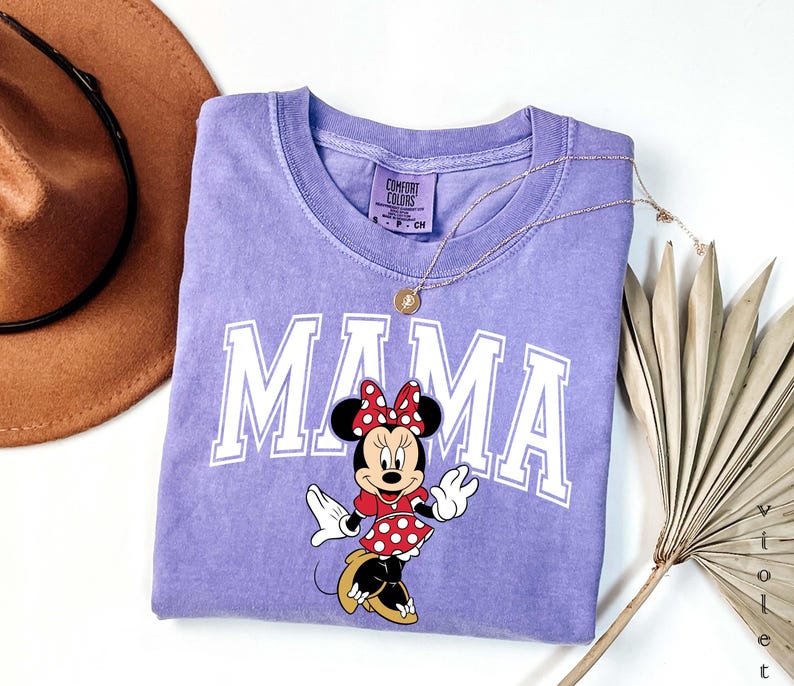 May include: A light purple t-shirt with the word "MAMA" in white block letters and a Minnie Mouse graphic. Minnie is wearing a red polka dot dress and bow. A gold necklace and a brown hat are also in the image.