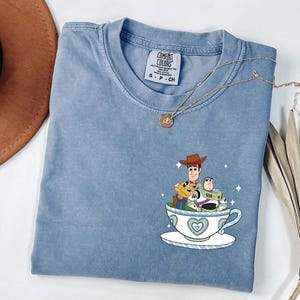 K&ouml;nnte beinhalten: Ein hellblaues Comfort Colors T-Shirt mit einer Grafik von Woody und Buzz Lightyear in einer Teetasse. Die Teetasse ist wei&szlig; mit einem blauen Herzdesign. Eine goldene Halskette ist &uuml;ber dem Shirt drapiert.