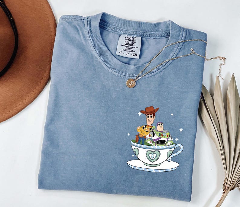 K&ouml;nnte beinhalten: Ein hellblaues Comfort Colors T-Shirt mit einer Grafik von Woody und Buzz Lightyear in einer Teetasse. Die Teetasse ist wei&szlig; mit einem blauen Herzdesign. Eine goldene Halskette ist &uuml;ber dem Shirt drapiert.