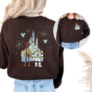 K&ouml;nnte beinhalten: Ein braunes Sweatshirt mit einem farbenfrohen Aufdruck von Woody, Buzz Lightyear und einem Schloss. Das Design umfasst Feuerwerk und Mickey Mouse-Ballons. Ein kleineres Bild zeigt die gleichen Charaktere in einer Tasse.