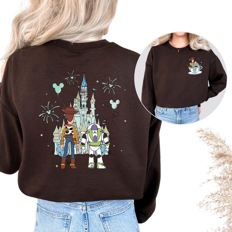 K&ouml;nnte beinhalten: Ein braunes Sweatshirt mit einem farbenfrohen Aufdruck von Woody, Buzz Lightyear und einem Schloss. Das Design umfasst Feuerwerk und Mickey Mouse-Ballons. Ein kleineres Bild zeigt die gleichen Charaktere in einer Tasse.