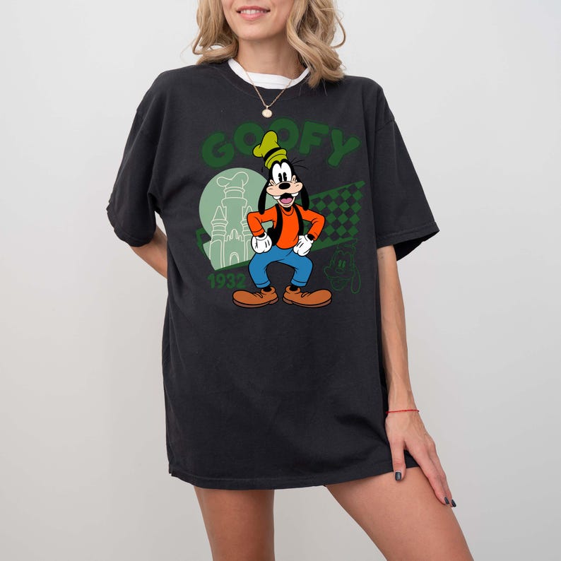 Może przedstawiać: Czarny t-shirt z zieloną grafiką Goofy'ego, postaci Disneya, oraz napisem "GOOFY". Projekt zawiera zamek, szachownicę i rok "1932". Koszulka ma kr&oacute;tkie rękawy i luźny kr&oacute;j.