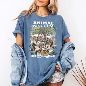 May include: Blue t-shirt featuring a graphic of Disney characters, including Mickey and Minnie Mouse, Donald Duck, and Goofy, with the text "ANIMAL KINGDOM" and "Walt Disney World". The shirt has rolled-up sleeves.