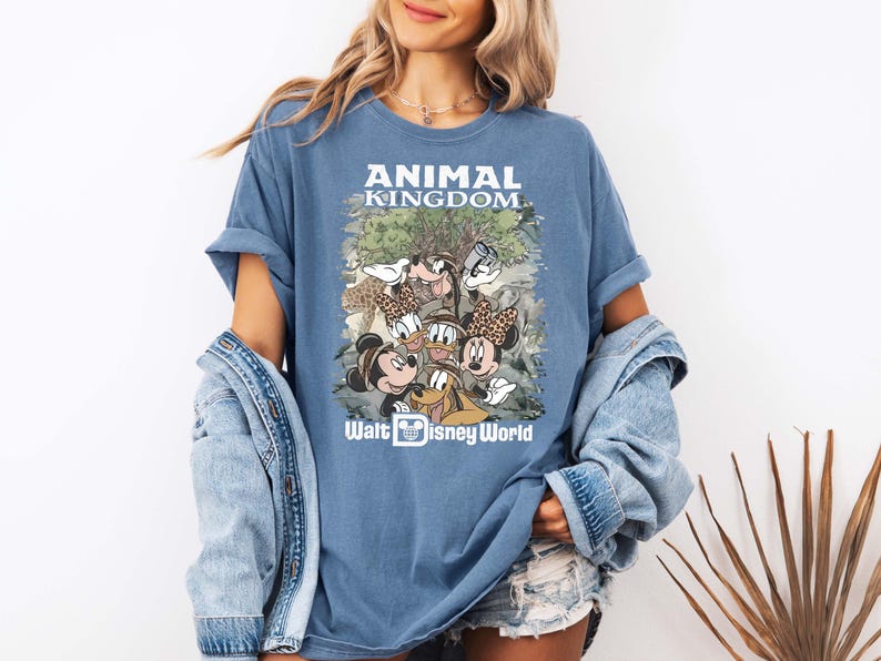 May include: Blue t-shirt featuring a graphic of Disney characters, including Mickey and Minnie Mouse, Donald Duck, and Goofy, with the text "ANIMAL KINGDOM" and "Walt Disney World". The shirt has rolled-up sleeves.