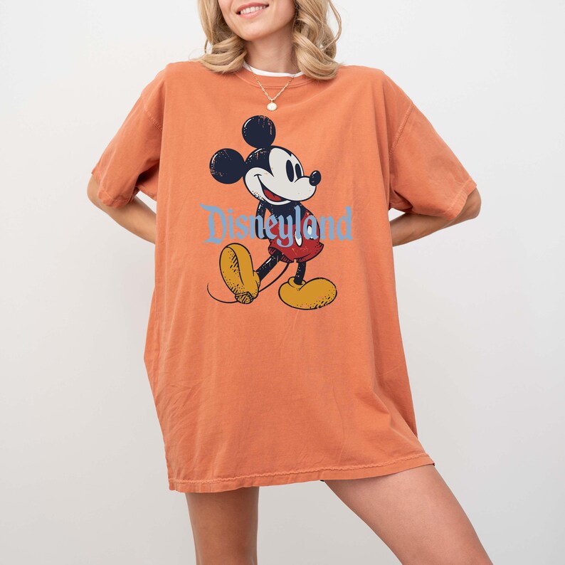 K&ouml;nnte beinhalten: Ein orangefarbenes T-Shirt mit einer Mickey Mouse-Grafik und dem Wort "Disneyland" in Hellblau. Mickey ist in seiner klassischen Pose dargestellt, mit schwarzen Ohren, einem wei&szlig;en Gesicht, roten Shorts und gelben Schuhen.