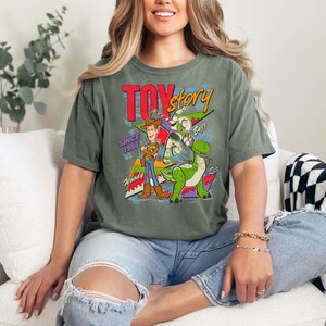 May include: Moss green t-shirt featuring a graphic from the movie Toy Story. The design includes Woody, Buzz Lightyear, and Rex, with the words "TOY STORY" and "SINCE 1995". The shirt is a relaxed fit.