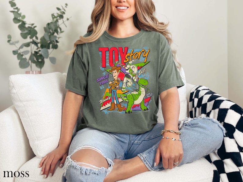 May include: Moss green t-shirt featuring a graphic from the movie Toy Story. The design includes Woody, Buzz Lightyear, and Rex, with the words "TOY STORY" and "SINCE 1995". The shirt is a relaxed fit.
