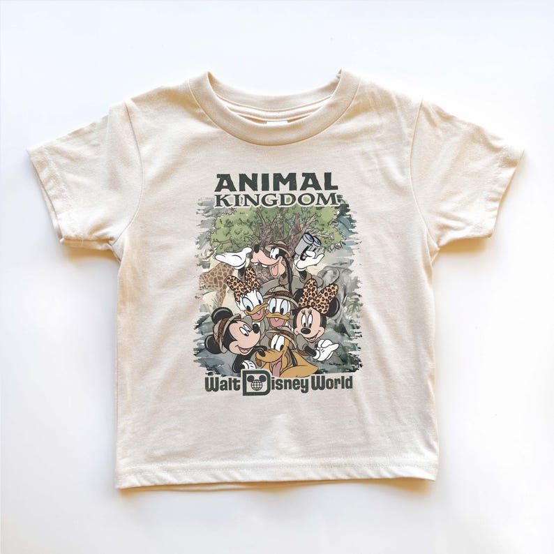 May include: Cream-colored t-shirt featuring a graphic of Disney characters, including Mickey and Minnie Mouse, Goofy, and Donald and Daisy Duck. The shirt has the text "ANIMAL KINGDOM" and "Walt Disney World" printed on it.