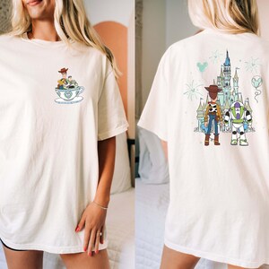 K&ouml;nnte beinhalten: Elfenbeinfarbenes T-Shirt mit Cartoon-Design. Auf der Vorderseite befindet sich eine kleine Grafik von Figuren in einer Teetasse. Auf der R&uuml;ckseite sind Woody und Buzz Lightyear vor einem Schloss mit Feuerwerk und einem Ballon abgebildet. Das Shirt hat eine lockere Passform.