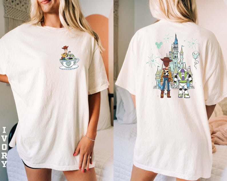 K&ouml;nnte beinhalten: Elfenbeinfarbenes T-Shirt mit Cartoon-Design. Auf der Vorderseite befindet sich eine kleine Grafik von Figuren in einer Teetasse. Auf der R&uuml;ckseite sind Woody und Buzz Lightyear vor einem Schloss mit Feuerwerk und einem Ballon abgebildet. Das Shirt hat eine lockere Passform.