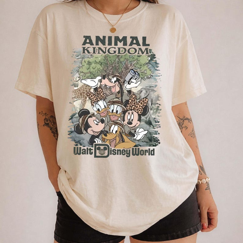 May include: Cream-colored t-shirt featuring a graphic of Disney characters, including Mickey and Minnie Mouse, Donald Duck, and Goofy, with the text "ANIMAL KINGDOM" and "Walt Disney World". The design has a vintage, cartoon style.