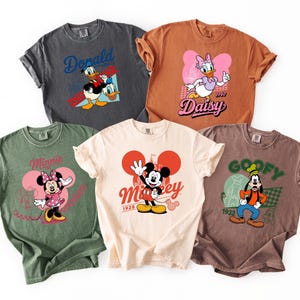 Może przedstawiać: Pięć kolorowych koszulek z postaciami Disneya. Koszulki są w r&oacute;żnych kolorach, w tym szarym, brązowym i zielonym. Każda koszulka ma inną grafikę postaci, taką jak Kaczor Donald, Daisy, Myszka Minnie, Myszka Miki i Goofy.