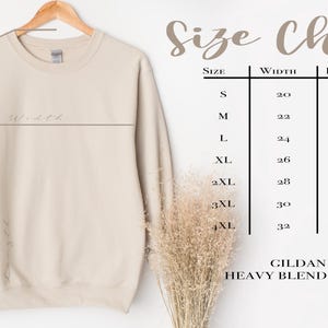 Może przedstawiać: Beżowa bluza z okrągłym dekoltem Gildan 18000 Heavy Blend wisi na drewnianym wieszaku. Tabela rozmiar&oacute;w pokazuje wymiary w cm dla szerokości, długości i długości rękawa, od rozmiaru S do 4XL.