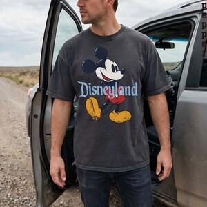 K&ouml;nnte beinhalten: Ein anthrazitfarbenes T-Shirt mit einem klassischen Mickey Mouse-Motiv und dem Wort "Disneyland" in blauer Schrift. Das Design zeigt Mickey's Gesicht, rote Shorts, gelbe Schuhe und schwarze Handschuhe. Das T-Shirt wird von einer Person getragen, die neben einem Auto steht.