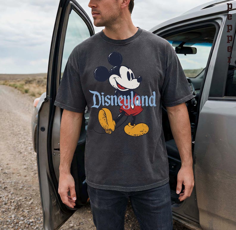 K&ouml;nnte beinhalten: Ein anthrazitfarbenes T-Shirt mit einem klassischen Mickey Mouse-Motiv und dem Wort "Disneyland" in blauer Schrift. Das Design zeigt Mickey's Gesicht, rote Shorts, gelbe Schuhe und schwarze Handschuhe. Das T-Shirt wird von einer Person getragen, die neben einem Auto steht.