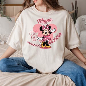 Może przedstawiać: Kremowa koszulka z grafiką Myszki Minnie w r&oacute;żowej sukience w kropki, kokardą i butami. Słowo "Minnie" znajduje się nad postacią, a pod nim "1928". R&oacute;żowe serce i wz&oacute;r w szachownicę dopełniają projekt.