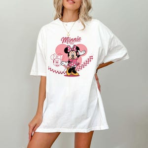 Może przedstawiać: Biała koszulka z grafiką Myszki Minnie. Projekt przedstawia Minnie w r&oacute;żowej sukience w kropki, r&oacute;żowej kokardce i napisem "Minnie" nad nią. Grafika umieszczona jest na r&oacute;żowym tle w kształcie serca.