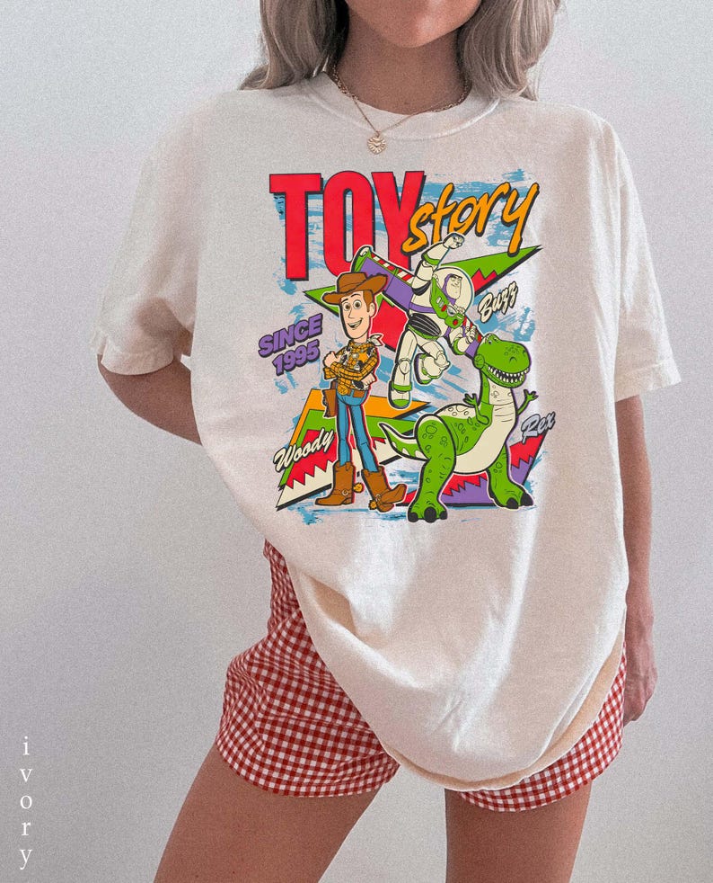 May include: Ivory-colored t-shirt featuring a graphic of Woody, Buzz Lightyear, and Rex from Toy Story. The design includes the text "Toy Story" in red, "Since 1995", and character names. The shirt is paired with red and white checkered shorts.