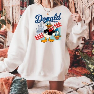Może przedstawiać: Biała bluza z nadrukiem Kaczora Donalda i napisem "Donald 1934". Projekt zawiera zamek i czerwono-biały wz&oacute;r w kratę. Swobodny, wygodny styl.