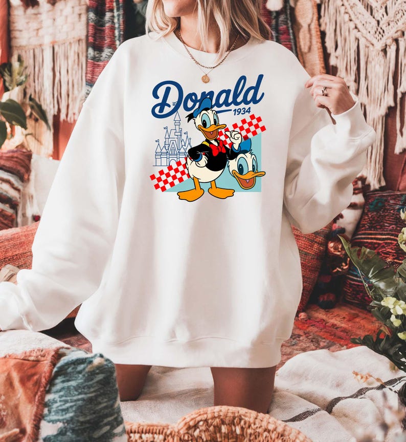 Może przedstawiać: Biała bluza z nadrukiem Kaczora Donalda i napisem "Donald 1934". Projekt zawiera zamek i czerwono-biały wz&oacute;r w kratę. Swobodny, wygodny styl.