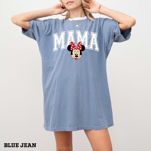 May include: Blue jean colored t-shirt with the word "MAMA" in white block letters and a Minnie Mouse graphic. The graphic features Minnie Mouse's face with a red bow. The shirt has a white collar.