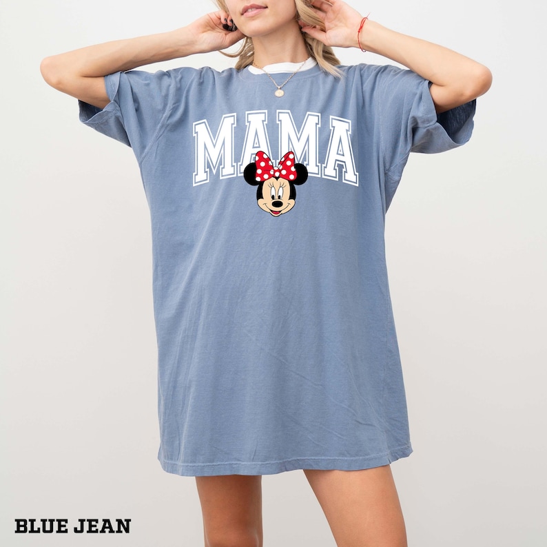 May include: Blue jean colored t-shirt with the word "MAMA" in white block letters and a Minnie Mouse graphic. The graphic features Minnie Mouse's face with a red bow. The shirt has a white collar.