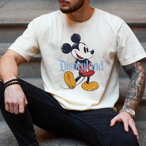 K&ouml;nnte beinhalten: Cremefarbenes T-Shirt mit einem Mickey Mouse-Motiv im Vintage-Stil und dem Wort "Disneyland" in Hellblau. Mickey wird in seiner klassischen Pose dargestellt, mit roter Hose und gelben Schuhen.