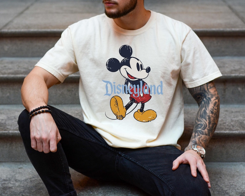 K&ouml;nnte beinhalten: Cremefarbenes T-Shirt mit einem Mickey Mouse-Motiv im Vintage-Stil und dem Wort "Disneyland" in Hellblau. Mickey wird in seiner klassischen Pose dargestellt, mit roter Hose und gelben Schuhen.